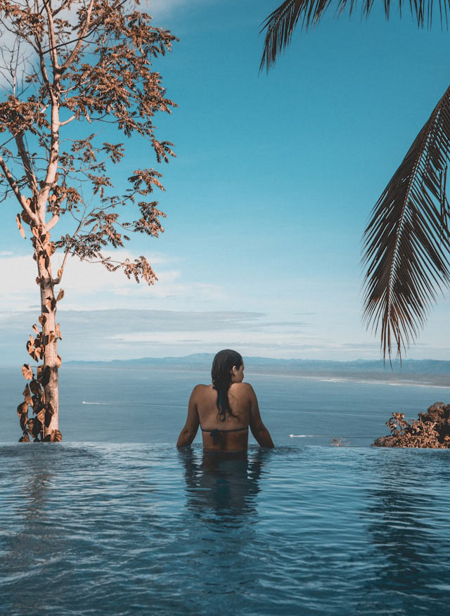Woman seen from the back, standing at the edge of an infinity pool, looking at the horizon, around her are trees and palms
